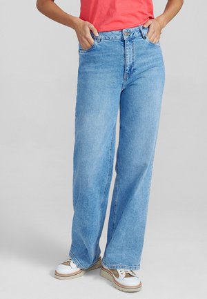 Persoon draagt lichtblauwe wijde jeans met handen in de zakken, witte en beige sneakers en een koraalkleurig kortemouwtop.