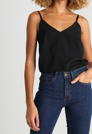 Vrouw draagt een zwarte mouwloze top met V-hals, ingestopt in een high-waisted donkerblauwe spijkerbroek, met een hand in de zak.