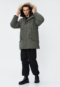 Olivgrüne Steppjacke mit einem mit Fell gefütterten Kapuzen, großen Fronttaschen und einem gesteppten Muster; kombiniert mit schwarzen Cargo-Hosen und Stiefeln.