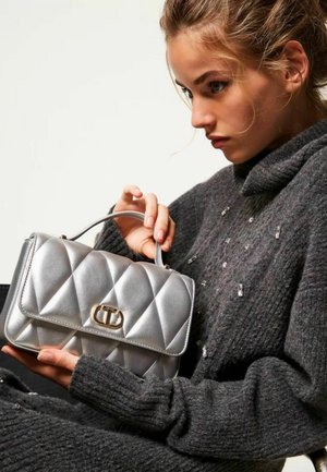Borsa a tracolla argentata trapuntata con una chiusura in metallo dorato, tenuta da una persona in un maglione scuro e strutturato. Materiale liscio e lucido e design ben definito.