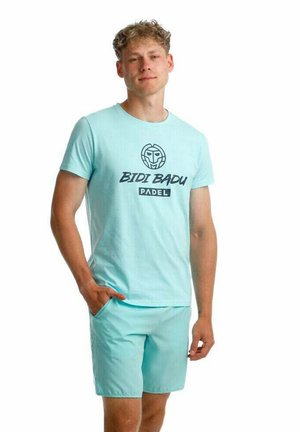 Joven con camiseta azul claro de Bidi Badu Padel y pantalones cortos a juego, de pie con una mano en el bolsillo.