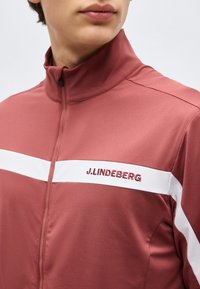 Κοντινό πλάνο ενός ατόμου που φοράει ροζ ζακέτα με φερμουάρ, με λευκή οριζόντια ρίγα και κέντημα "J. LINDEBERG" στο στήθος.