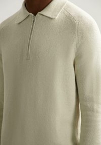 Svijetlosmeđi pleteni pulover s ovratnikom na zip. Ima mekan, teksturiran površinu i prilagođen kroj s raglan rukavima.