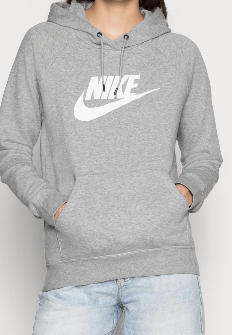 Personne portant un sweat à capuche gris Nike avec logo blanc et un jean bleu clair, les mains dans les poches du sweat, sur un fond uni.