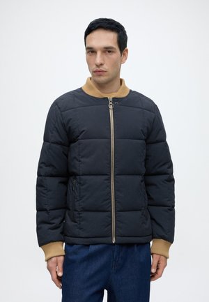 Homme portant une veste matelassée bleu marine avec col et poignets côtelés beige, debout les mains détendues le long du corps devant un fond clair uni.