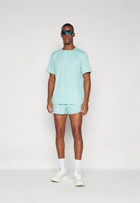 Férfi áll, világoskék sportnadrágban és pólóban, fehér zokniban, fehér futócipőben és fényvisszaverő sportnapernyőben, egyszínű háttér előtt.