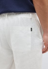 Personne portant un pantalon en lin blanc avec une poche arrière ornée d'une petite étiquette noire de la marque Ecoalf, associée à un haut bleu foncé.