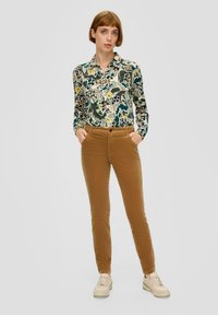Vrouw staat met haar handen in haar zakken, gekleed in een bloemenoverhemd, bruine corduroy broek en lichtbeige sneakers.
