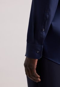 Camicia in satin blu navy con polsino a punta e bottone. Tessuto liscio e vestibilità su misura, con dettagli sottili vicino al polsino.