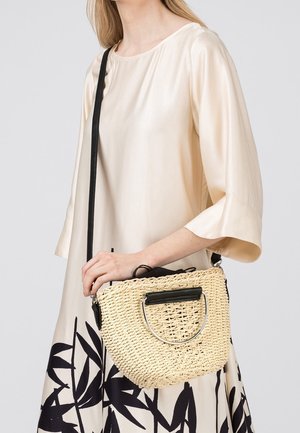 Vrouw draagt een crèmekleurige jurk met een zwart bladmotief, houdt een gevlochten strooien crossbodytas vast met zwarte en zilveren handvatten.