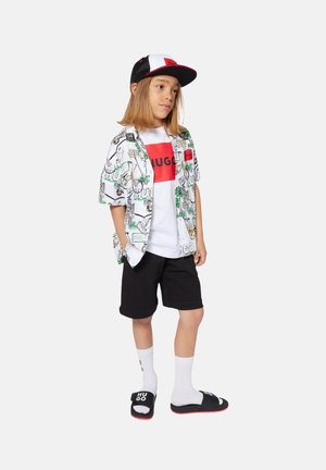 Kortebloezem witte shirt met palmbomenprint, rood logo t-shirt, zwarte shorts, witte sokken en zwarte slipper sandalen met logo details.