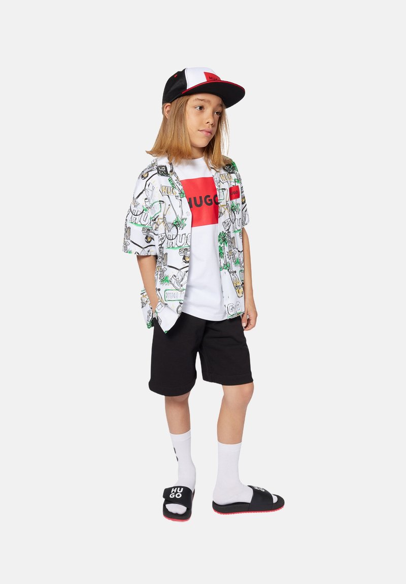 Kortebloezem witte shirt met palmbomenprint, rood logo t-shirt, zwarte shorts, witte sokken en zwarte slipper sandalen met logo details.
