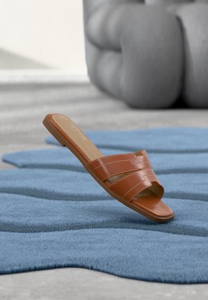 Sandale mule en cuir brun posée sur un tapis bleu texturé ondulé avec un fauteuil gris flou en arrière-plan.