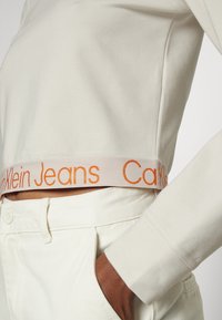 Skórzany, krótki sweatshirt w kolorze ecru, z widocznym, pomarańczowym, haftowanym logo na elastycznym pasku, połączony z białymi spodniami.