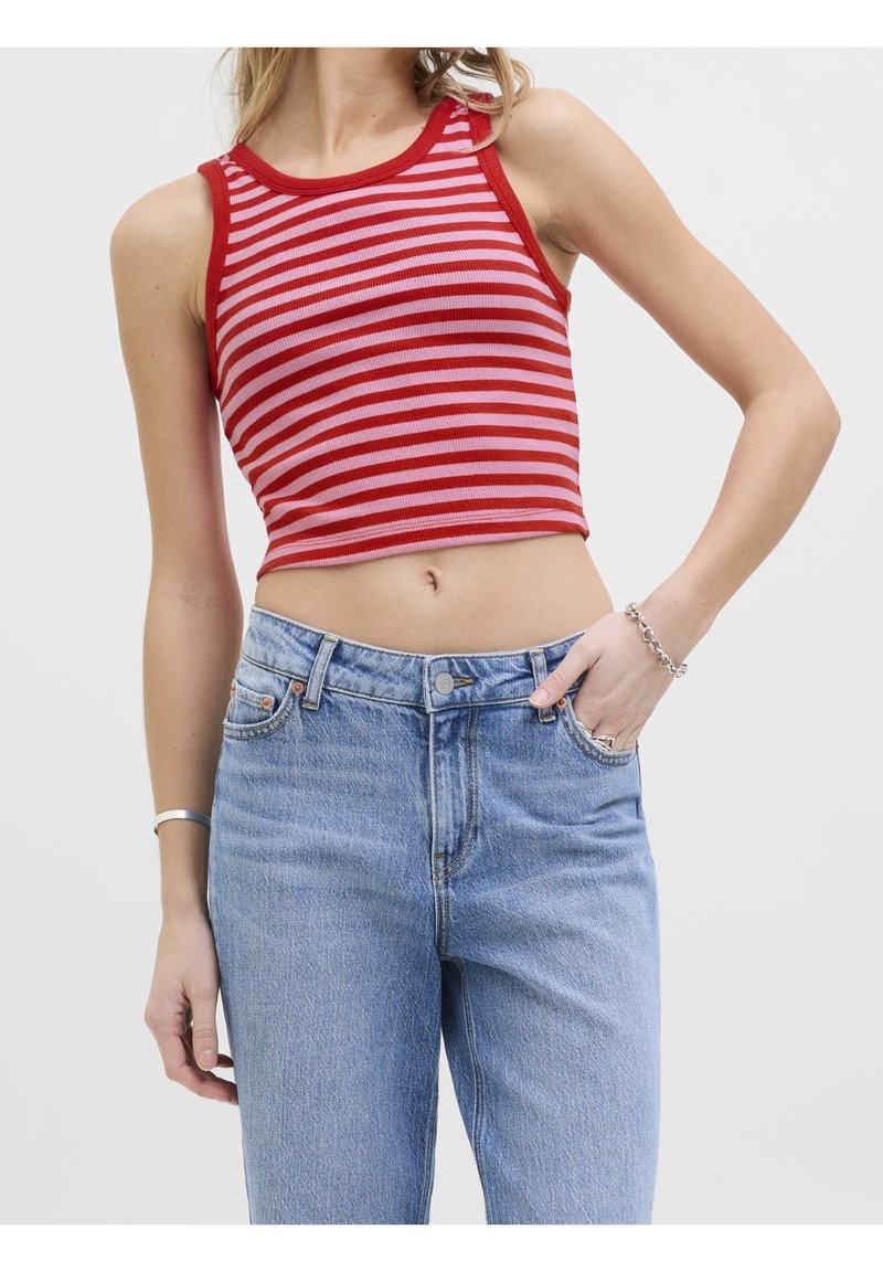 Femme portant un top court sans manches rayé rouge et rose et un jean bleu clair taille haute, une main dans la poche et des bracelets argentés.