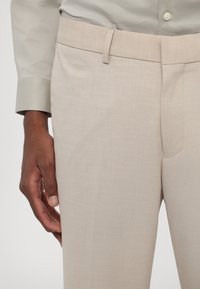 Pantaloni beige su misura con una superficie liscia, caratterizzati da un design a vita piatta e un sottile motivo a tessitura. La mano è appoggiata sul tessuto.