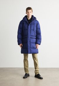 Cappotto imbottito blu con cappuccio, dotato di chiusura con zip e bottoni a pressione, e due tasche frontali. Indossato sopra una camicia nera con colletto e pantaloni khaki.
