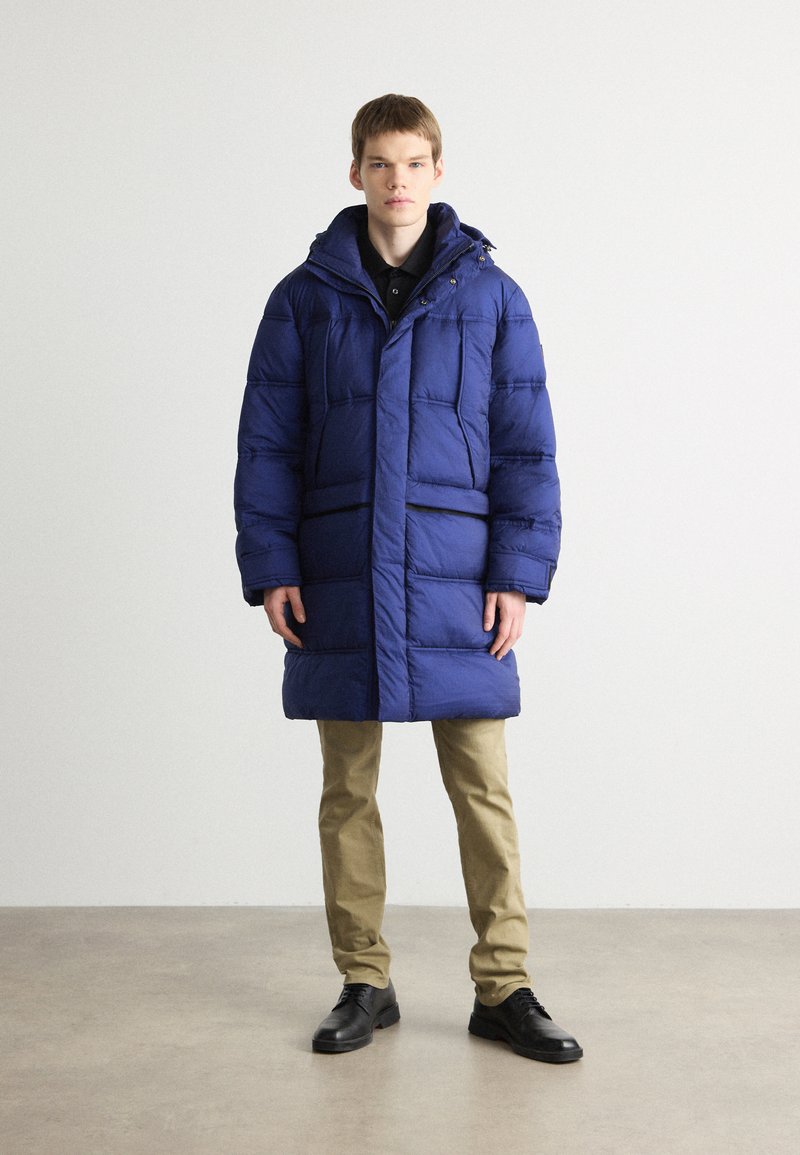 Cappotto imbottito blu con cappuccio, dotato di chiusura con zip e bottoni a pressione, e due tasche frontali. Indossato sopra una camicia nera con colletto e pantaloni khaki.
