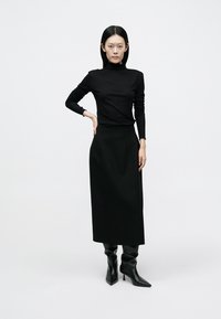 Moteris su tiesiais juodais plaukais, dėvinti juodą golfą, ilgą juodą sijoną ir juodus smailius kulkšnies aulinukus, stovinti viena ranka ant klubo.