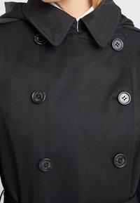 Svart trenchcoat med dubbelknapprad, stor krage och mörka knappar. Tillverkad av ett mjukt tyg med subtil textur och sömnadsdetaljer.