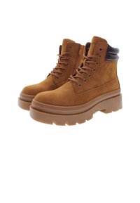 Botas de tobillo de ante marrón con un collarín acolchado negro, parte delantera con cordones y suela gruesa de goma texturizada. Presenta detalles del logo en el costado.