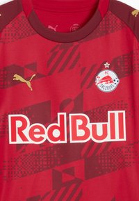 Camiseta roja con patrones texturizados en rojo más oscuro, con el logo dorado de Puma en el hombro, gran texto "Red Bull" y el emblema del FC Salzburg.