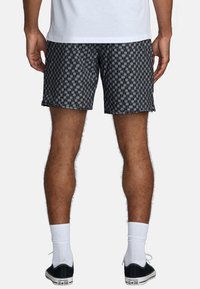 Zwarte zwembroek met een geometrisch patroon, met een lengte tot halverwege de dijen en een ontspannen pasvorm. G gedragen met witte sokken en zwarte sneakers.