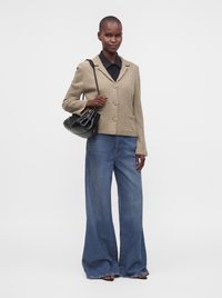 Femme portant un cardigan beige en maille torsadée, une chemise noire, un jean bleu à jambes larges, et tenant un sac à main noir sur son épaule.