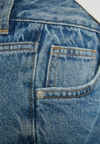 Los jeans de mezclilla de lavado claro cuentan con dos bolsillos traseros, costuras visibles en color naranja y un detalle de botón metálico en el bolsillo lateral.