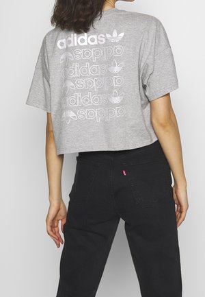 Une personne porte un haut gris Adidas raccourci avec un logo répété au dos et un jean noir taille haute avec les poches arrière visibles.