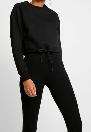 Frau trägt ein schwarzes, kurzes Sweatshirt mit Fronttasche und Kordelzug, kombiniert mit schwarzen Leggings mit einem dezenten Seitenmuster.