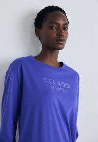 Camisa de mangas compridas roxa, feita de tecido suave, apresentando um gráfico das fases da lua e o texto "à luz da lua" em branco.