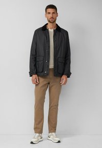 Mann steht an einer grauen Wand und trägt eine schwarze Jacke mit großen Taschen, einen beigen Pullover, braune Hosen und weiße Sneakers mit beigen Akzenten.