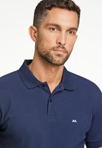 Marinblå polo skjorta gjord av strukturerad väv, med en treknappslucka och en liten bergslogotyp på vänster bröst.