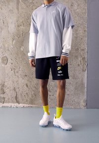 Light blue polo shirt with white long sleeves, navy shorts with logo prints, yellow socks, and white sneakers with transparent soles.