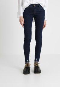 Jeans ajustados en denim azul oscuro con dobladillo desgastado. Combinados con zapatos de plataforma negros y una camisa blanca de botones.