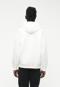 Sudadera blanca con capucha, corte holgado y hombros caídos. Terminada con puños y dobladillo acanalados. Textura suave, diseño minimalista.