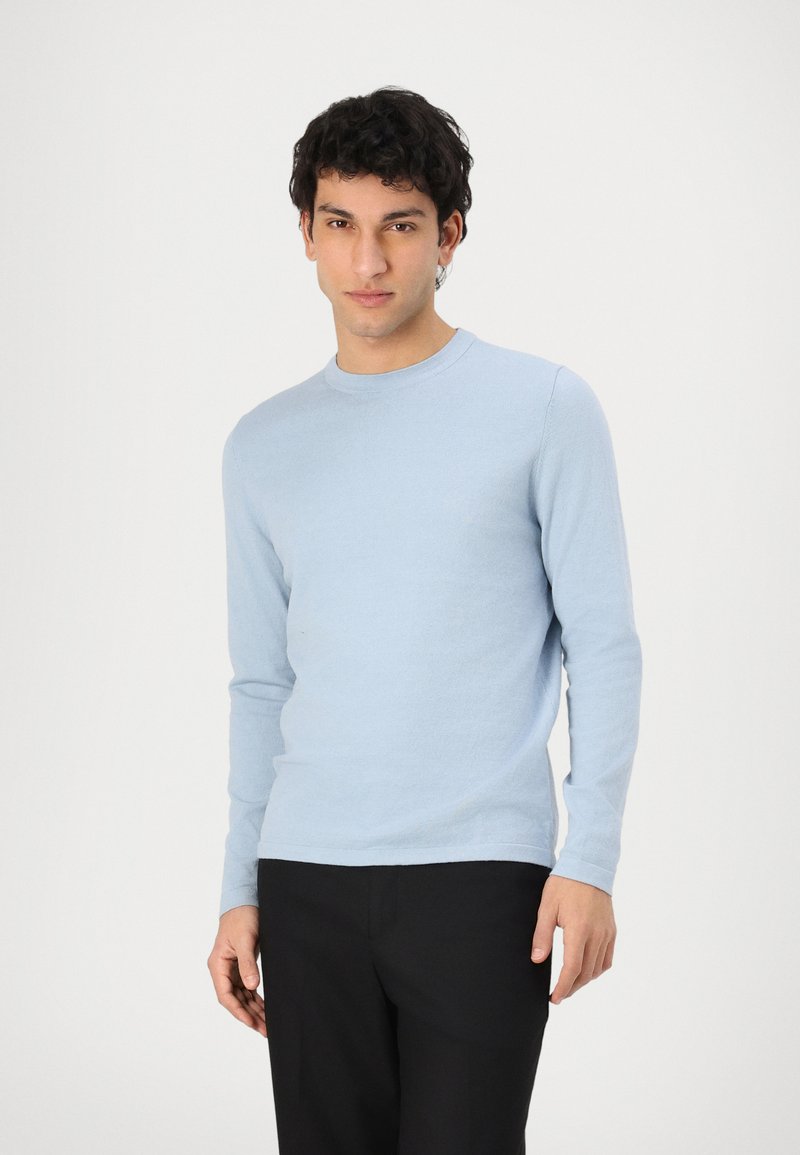 Hellblauer langärmeliger Pullover aus weichem Strickmaterial, mit Rundhalsausschnitt und figurnaher Passform, kombiniert mit schwarzen Hosen.