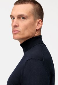 Maglione a collo alto blu navy realizzato in un tessuto a maglia testurizzato. Presenta un design aderente con collo a costine, evidenziando un dettaglio spalla senza cuciture.
