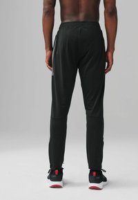Black athletic pants with a slim fit, elastic waistband, and side pockets. Features panel accents and is paired with black sneakers.