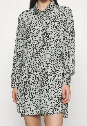 Robe à manches longues boutonnée avec un motif tacheté noir et blanc, portée par une personne avec les mains et les jambes visibles.
