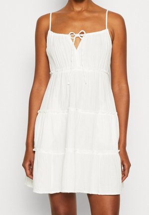 Kobieta w białej, warstwowej, mini sukience na ramiączkach z cienkimi paskami i kokardą z przodu przy dekolcie.