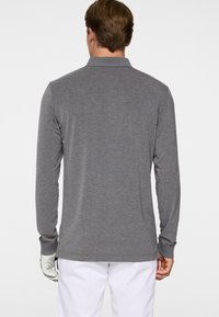 Mężczyzna ubrany w szarą bluzkę polo z długim rękawem i białe spodnie, zwrócony tyłem, z białą rękawiczką na lewej ręce.