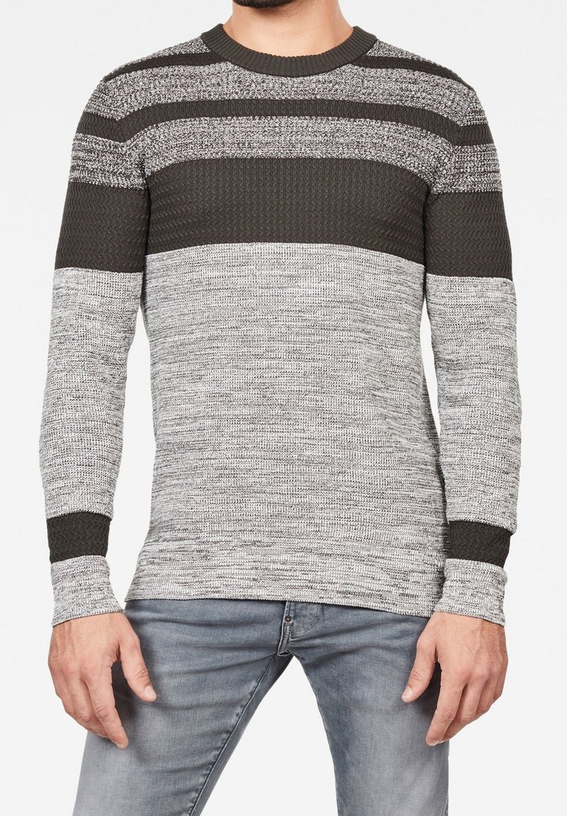 Homme portant un pull en maille gris et noir à rayures horizontales et un jean gris clair, debout devant un fond uni.
