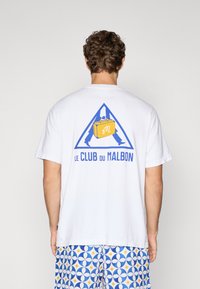 Hvid bomulds t-shirt med en blå trekantet logo med en gul kuffert og teksten "LE CLUB DU MALBON" på ryggen. Korte ærmer.