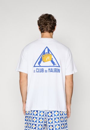 Hvit bomull t-skjorte med en blå trekantet logo som har en gul koffert og teksten "LE CLUB DU MALBON" på baksiden. Korte ermer.