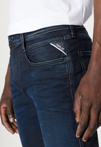 Dark blue denim jeans with a slim fit. Features a subtle "REPLAY" label on front pocket and classic pocket styling. Smooth texture.