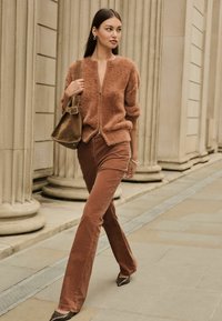 Un pull zippé du couleur rouille, duveteux, associé à un pantalon évasé assorti et des talons à motifs ; tenant un sac à bandoulière marron.