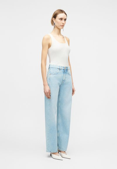 Femme portant un haut blanc sans manches à côtes, un jean taille haute bleu clair à jambes larges, des talons blancs pointus et des boucles d'oreilles remarquables.