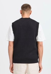Jonge man met kort haar die wegkijkt, in een zwart mouwloos brei-vest over een wit kortebloes met lichte kleurige broek.
